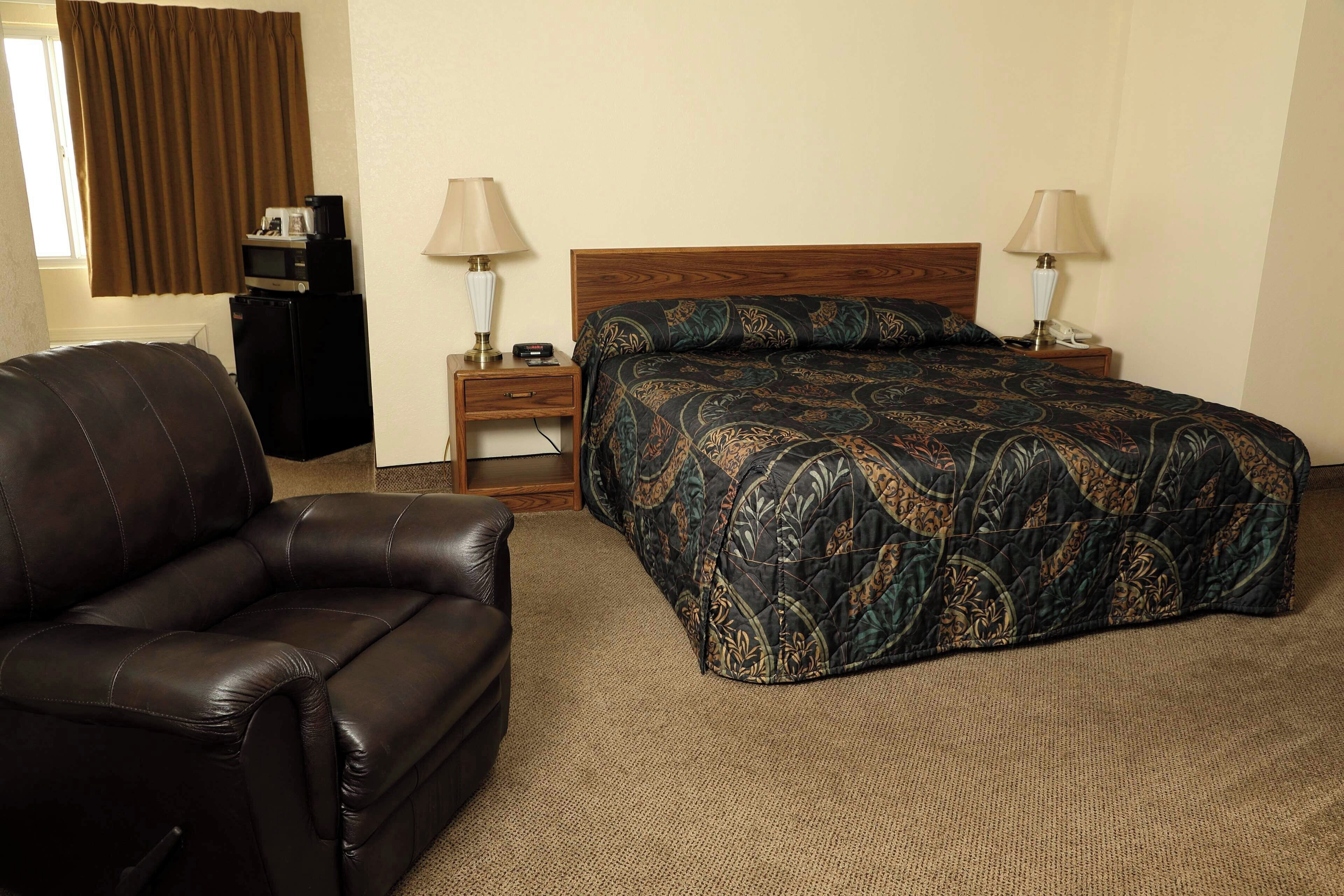 Une chambre d’hôtel comprenant un lit, un fauteuil en cuir, une table de chevet avec une lampe, un bureau avec une lampe, une télévision et une fenêtre avec des rideaux.