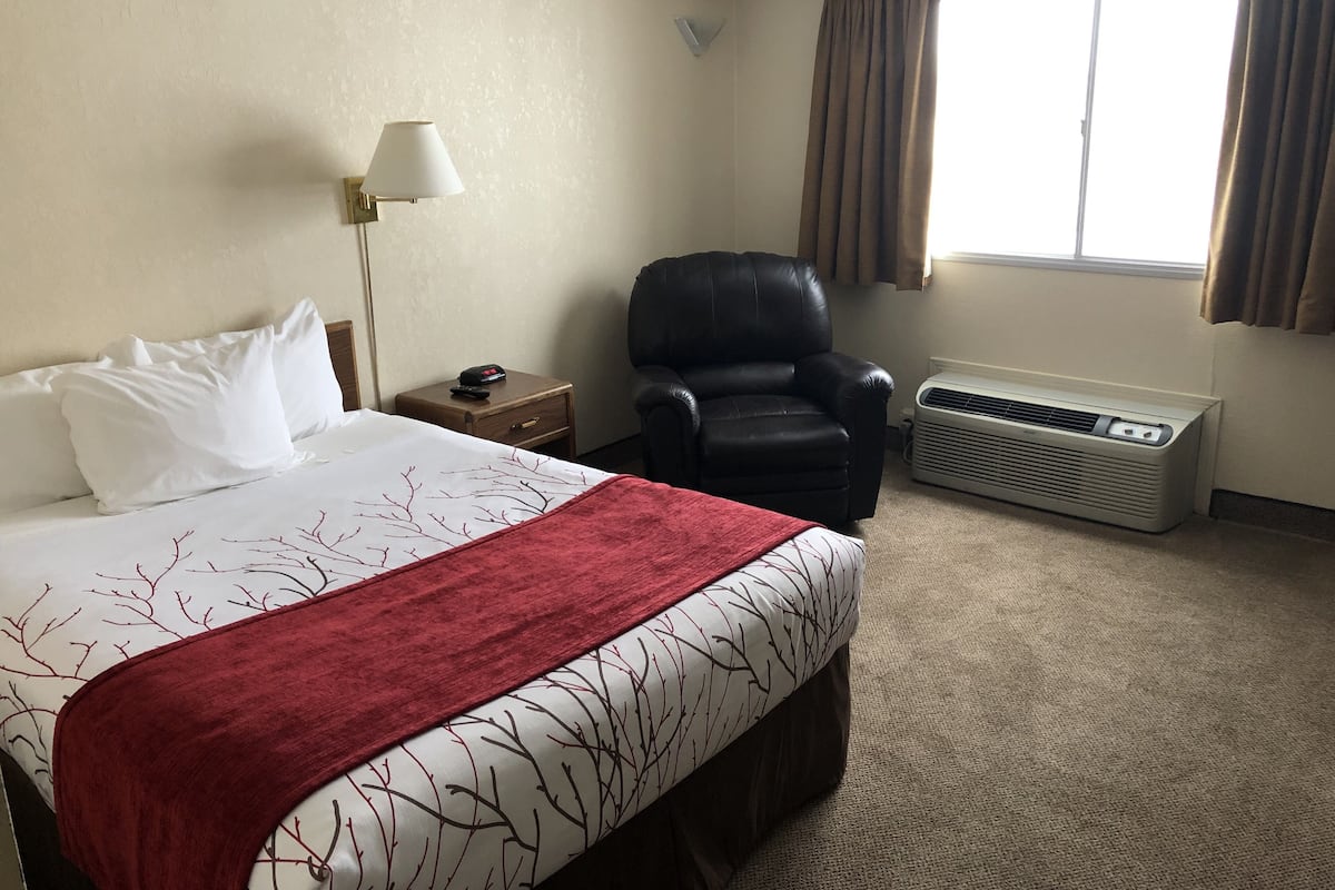 A hotel room with a bed, a chair, a nightstand, a lamp, an air conditioner, and a window with curtains.