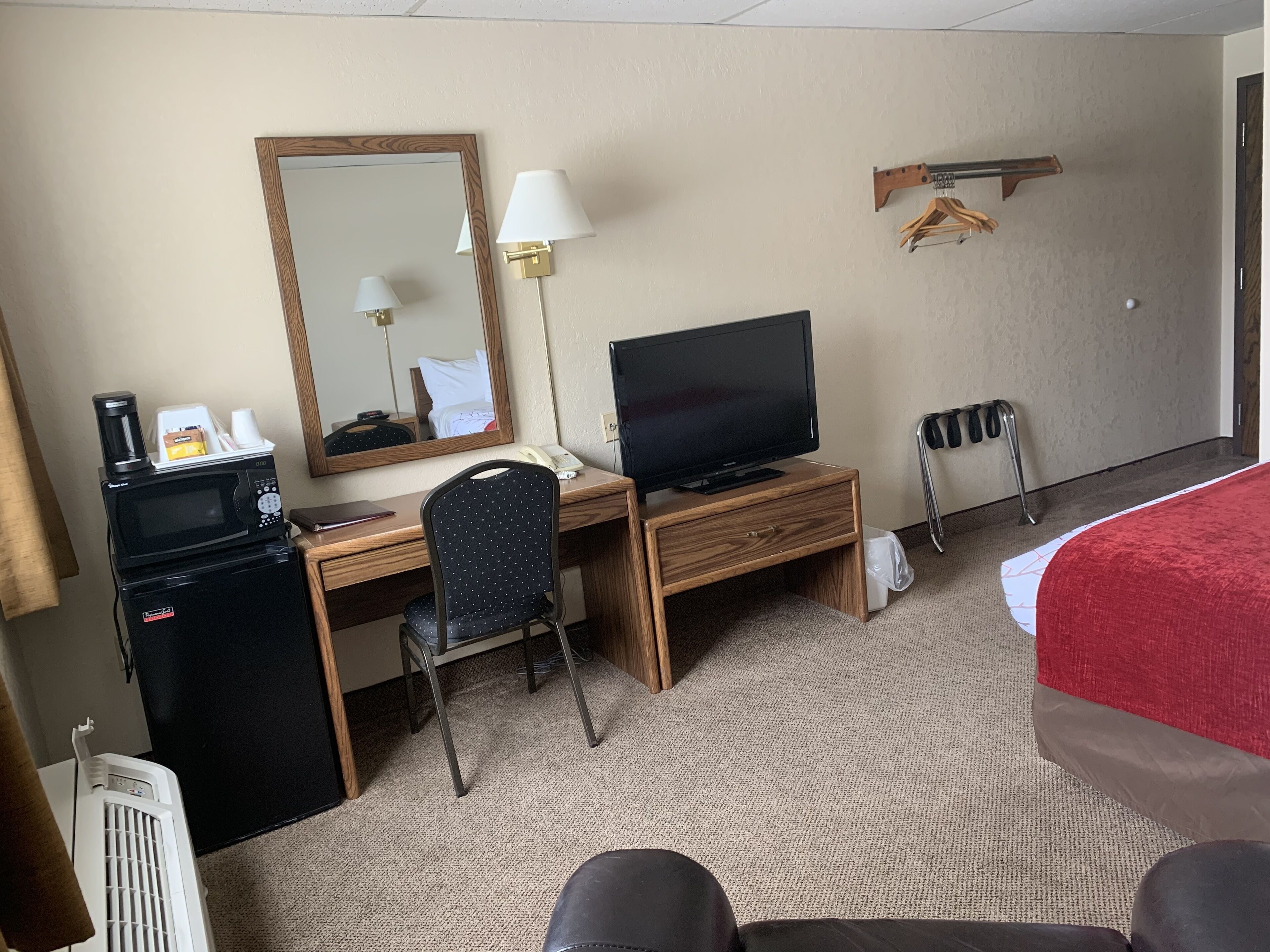 A hotel room with a wooden desk, a television, a chair, a microwave, a heater, and a bed with a red cover.