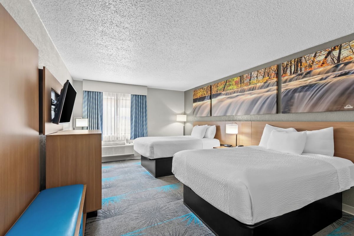 A hotel room with two beds, a desk, a TV, and a large painting of a waterfall.
