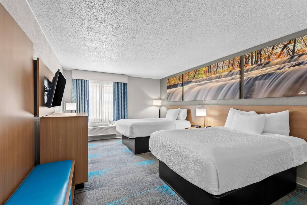 A hotel room with two beds, a desk, a TV, and a painting of a waterfall.