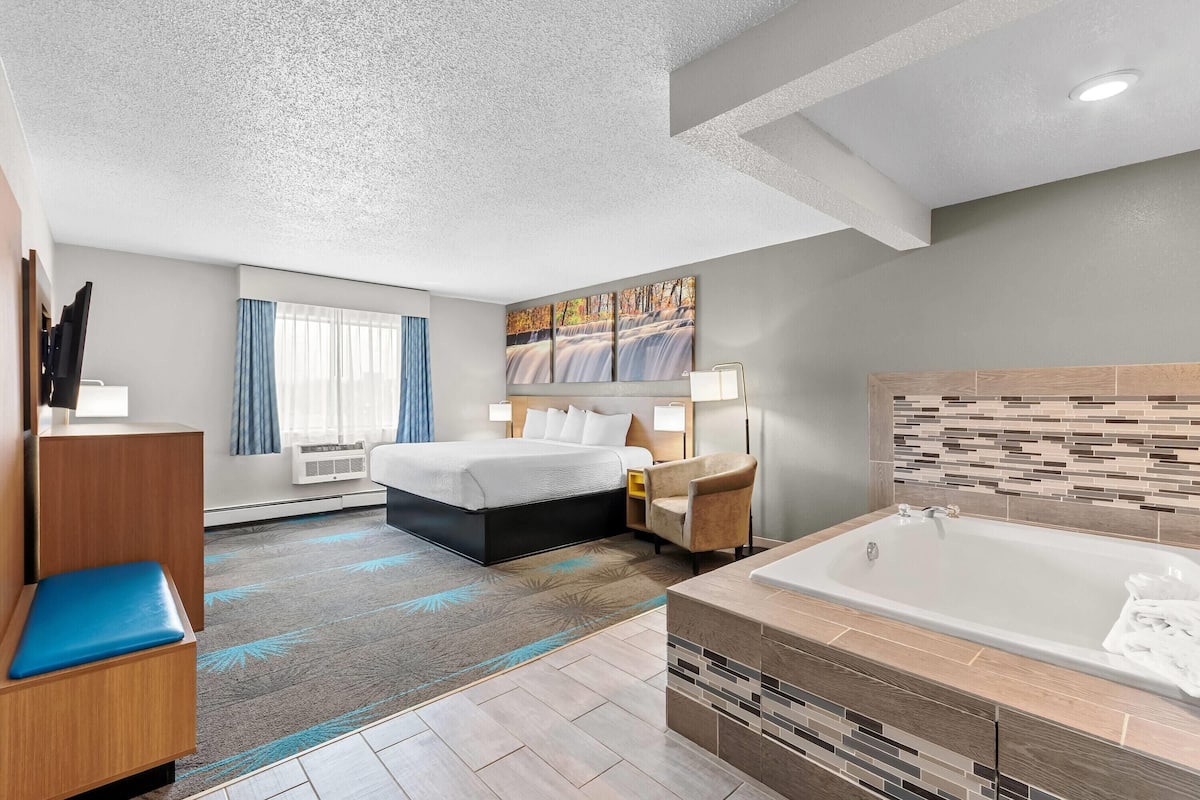 A hotel room with a bed, a desk, a chair, a bathroom with a bathtub, and a large window.