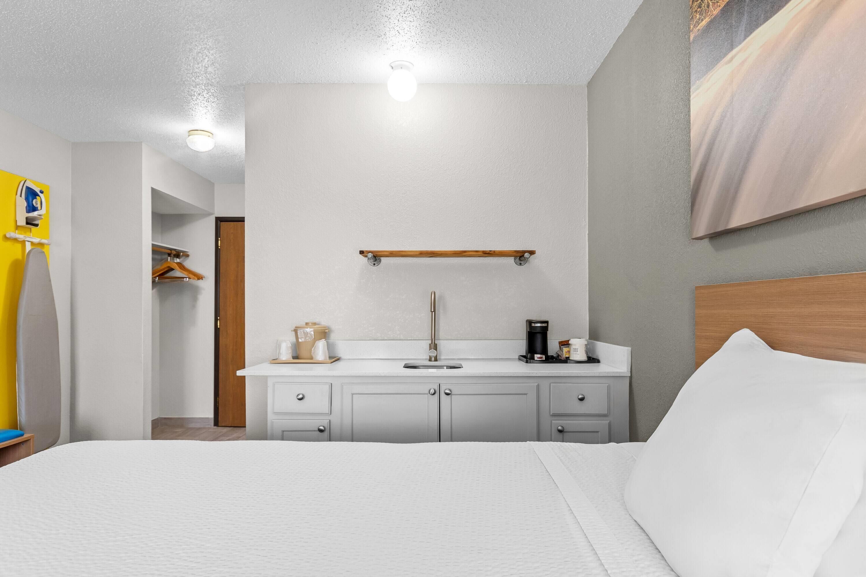 A hotel room with a bed, a white wall, a wooden headboard, a white bathroom vanity, a mirror, a coffee maker, and a painting.