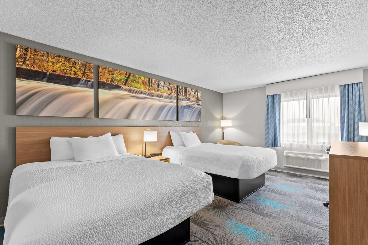 A hotel room with two beds, a large wall-mounted picture, a window with curtains, and a bedside table with a lamp.
