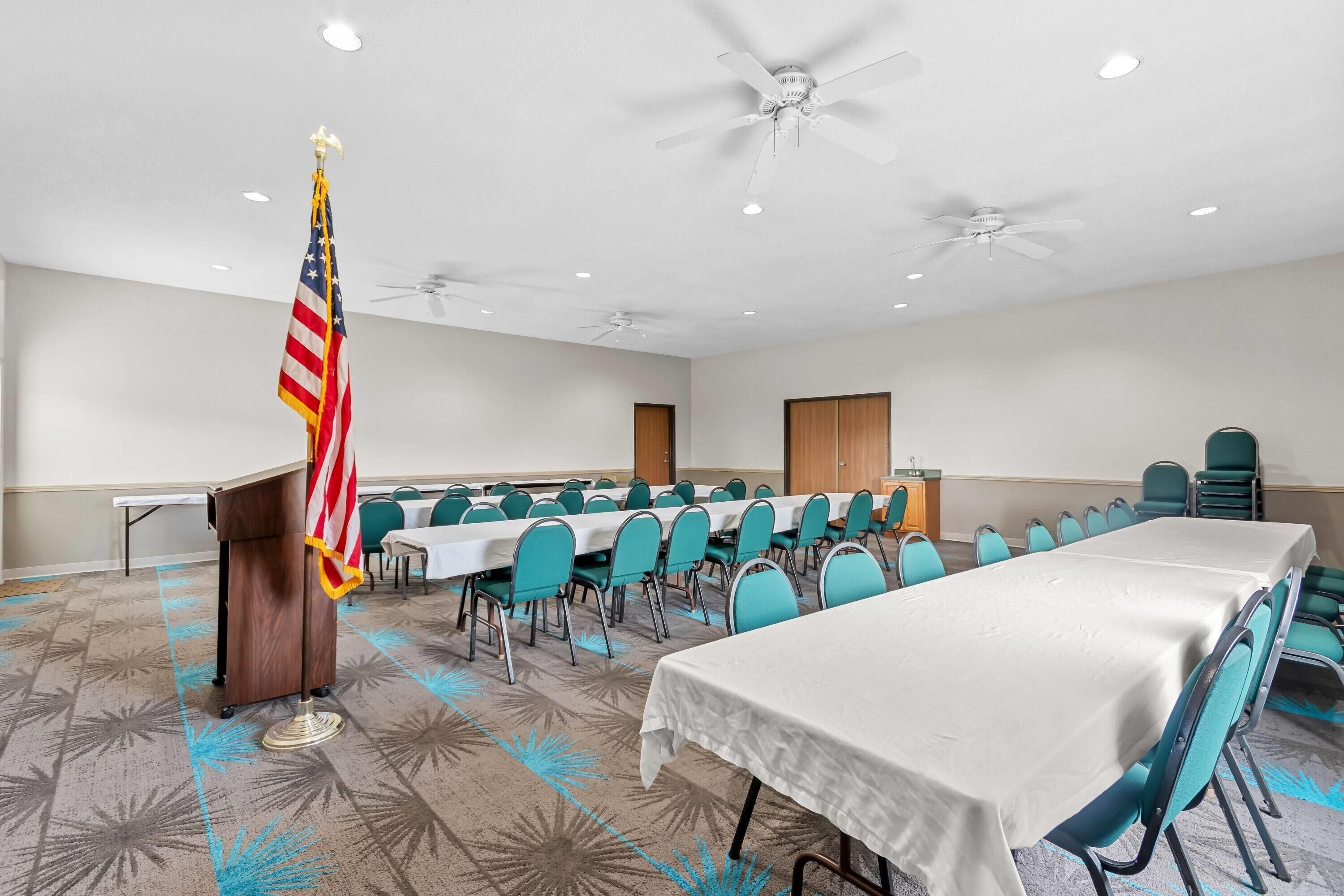 A conference room with a long table, teal chairs, a podium with an American flag, and ceiling fans.