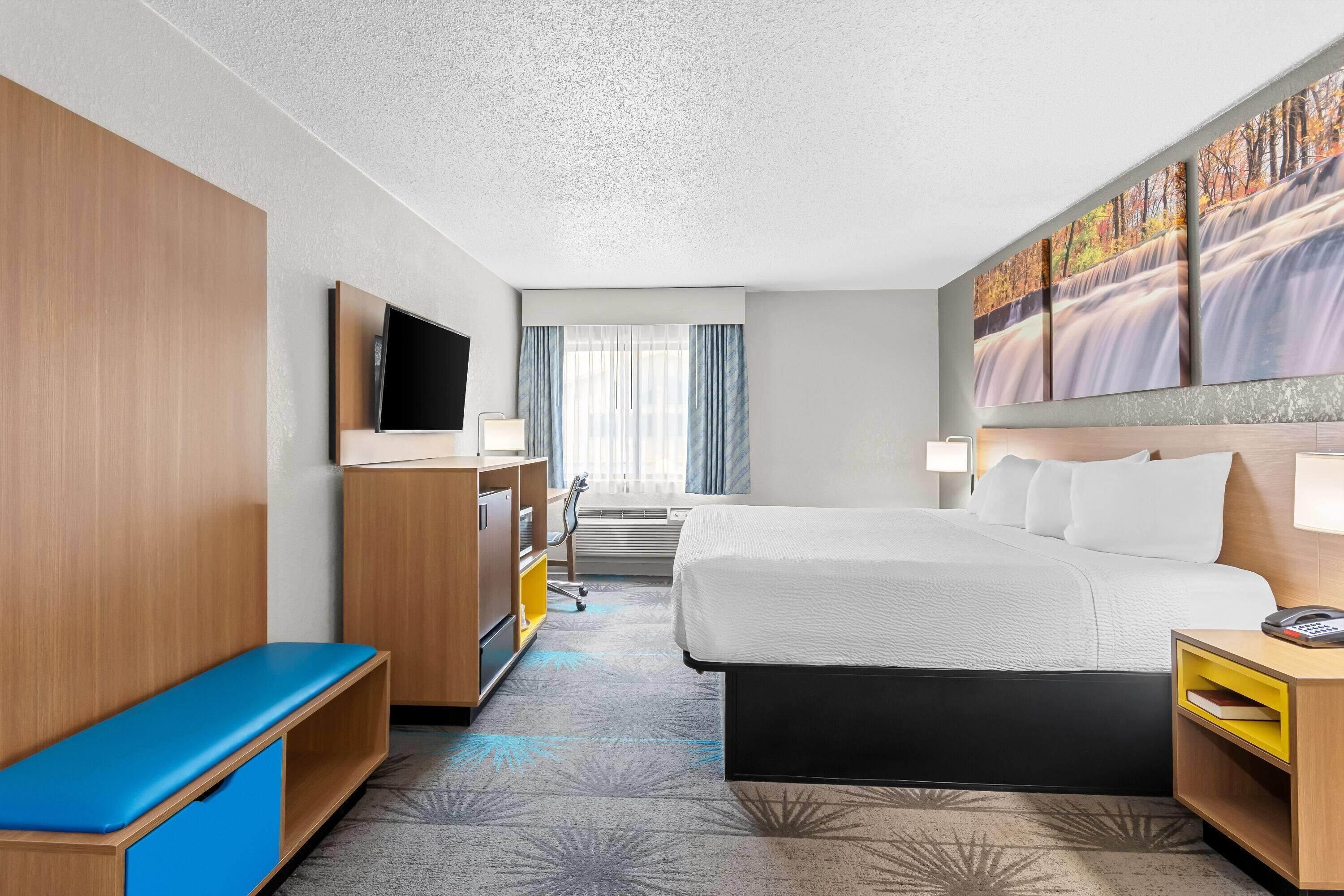 Habitación de hotel con cama, escritorio, televisión, ventana con cortinas y un cuadro en la pared.