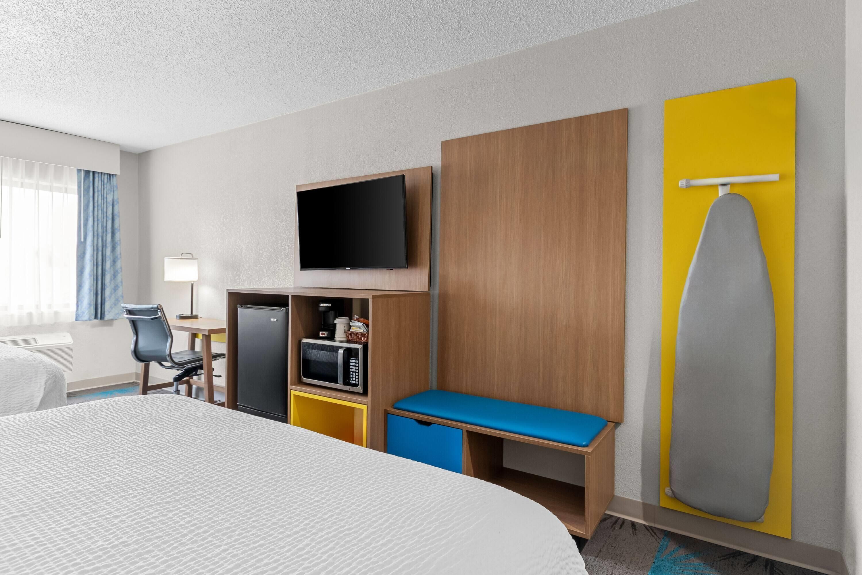 A hotel room with a bed, a TV, a microwave, a desk, a chair, and a bench.