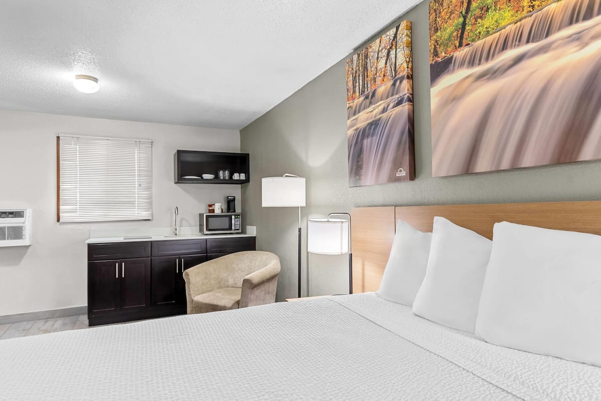 A hotel room with a bed, a desk with a chair, a microwave, a lamp, and artwork on the wall.