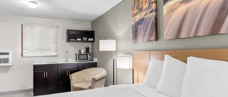 A hotel room with a bed, a desk with a chair, a microwave, a lamp, and artwork on the wall.