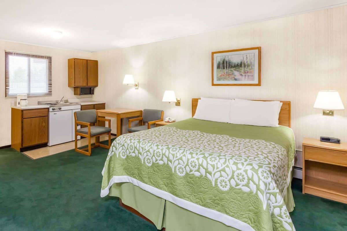 A hotel room with a bed, a desk with a chair, a kitchenette, and a painting on the wall.