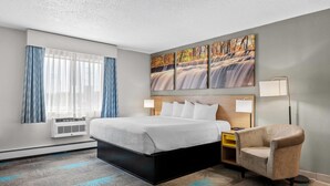 A hotel room with a bed, a chair, a desk, a lamp, and a wall-mounted air conditioner. - Days Inn by Wyndham Alpena (Alpena)
