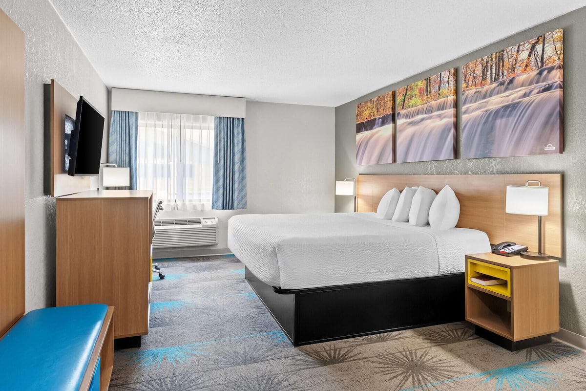 A hotel room with a bed, bedside tables, a desk, a TV, and a large window with curtains.