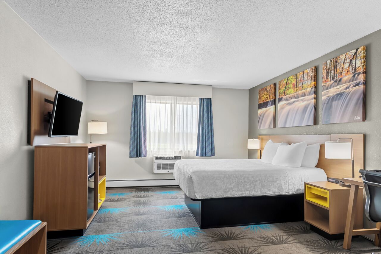 A hotel room with a bed, a desk, a TV, a window with curtains, and artwork on the wall.
