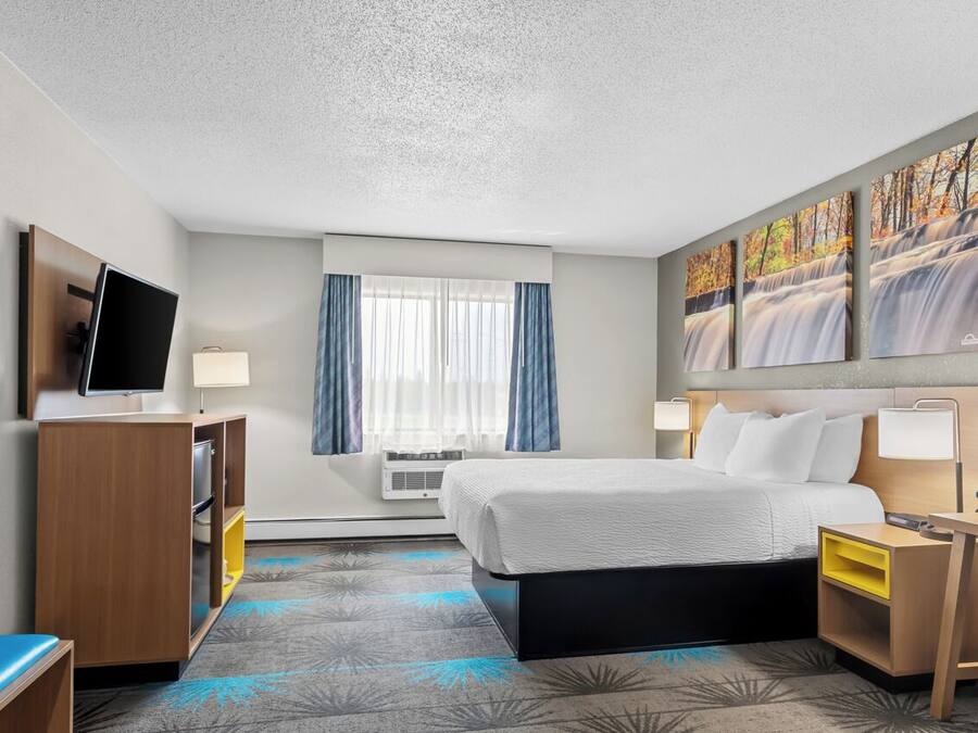 A hotel room with a bed, a desk, a TV, a window with curtains, and artwork on the wall.