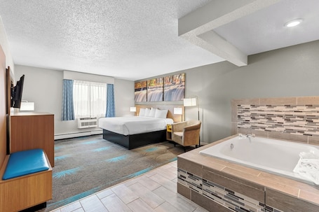 A hotel room with a bed, a desk, a chair, a large window with curtains, a painting, a bathtub, and a tiled wall.