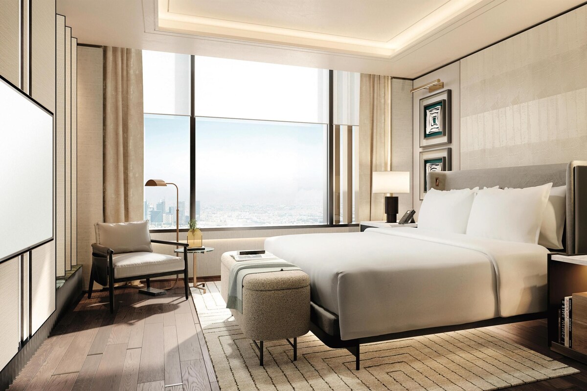Ein Hotelzimmer mit einem großen Bett, einer Sitzecke mit Sessel und kleinem Tisch, einem Nachttisch mit Lampe und Blick auf die Stadt durch große Fenster.