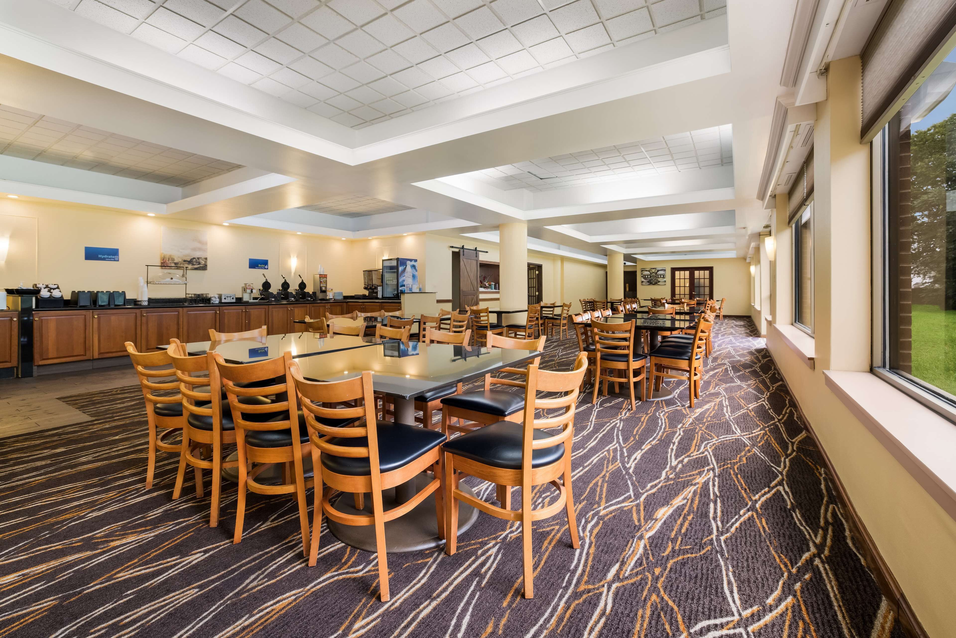 A spacious hotel dining area with multiple tables and chairs, a kitchen area, and large windows providing natural light.