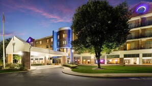 Um hotel de vários andares com design moderno, apresentando uma luminária circular destacada na fachada, um gramado bem cuidado e uma área de estacionamento.