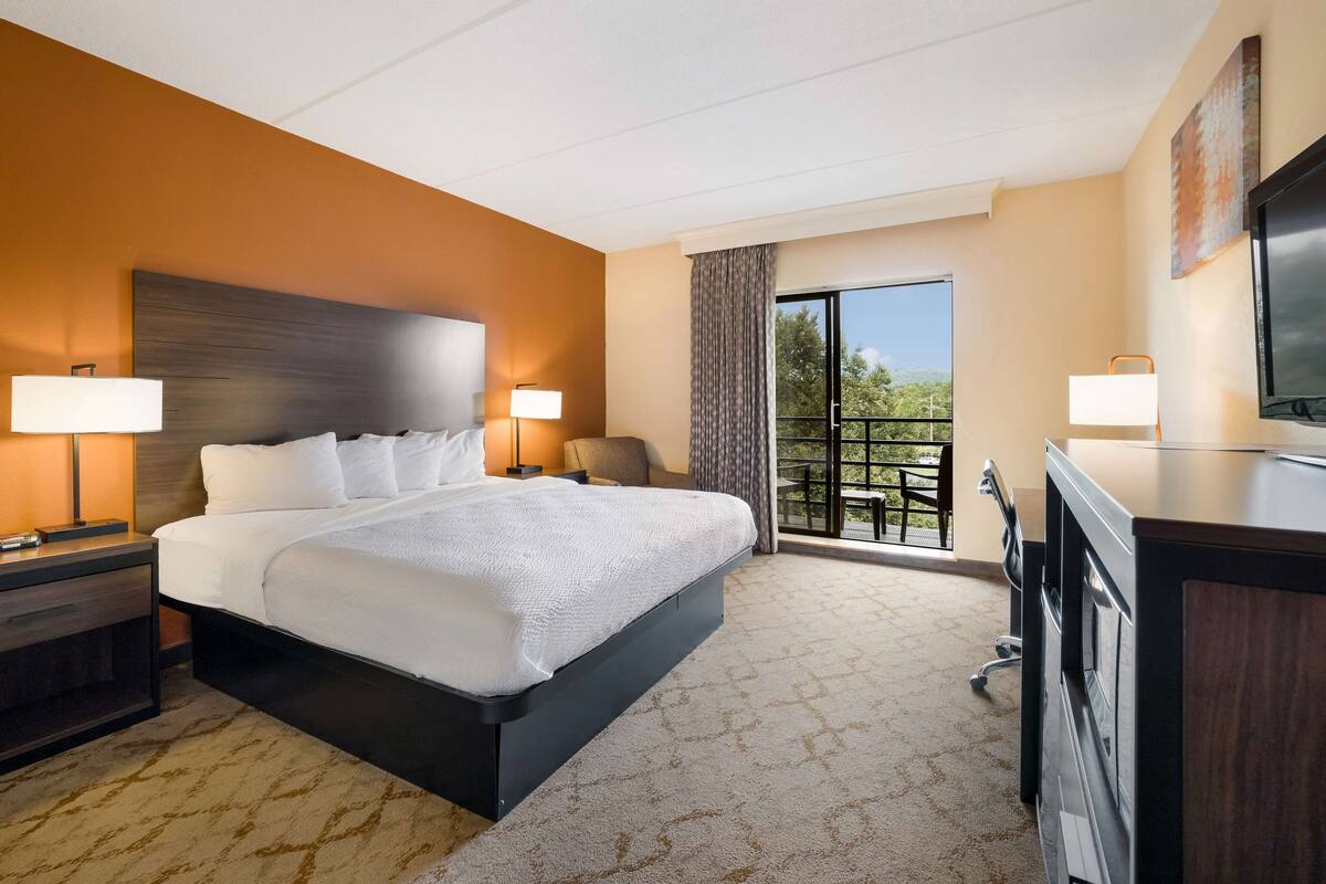 A hotel room with a large bed, two bedside lamps, a desk with a TV, and a balcony with a view.