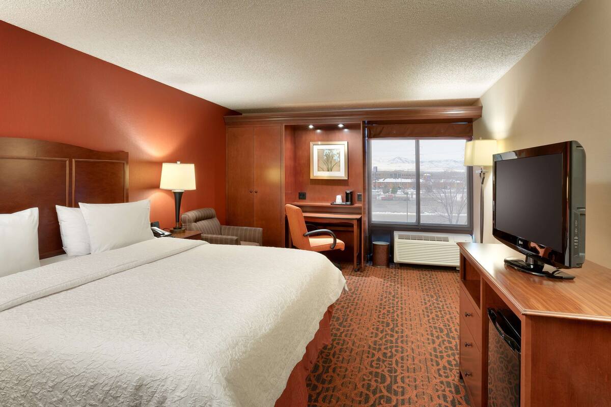 A hotel room with a large bed, a desk with a chair, a television, and a window with a view of a building and mountains.