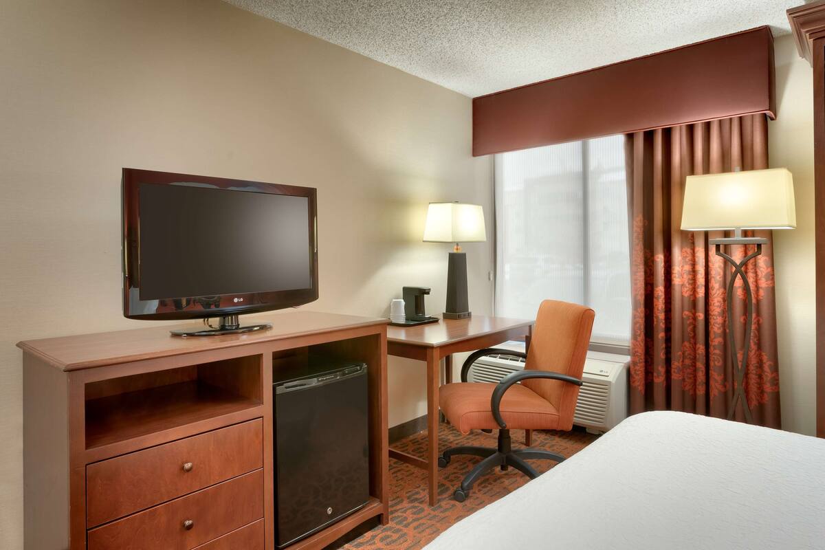 A hotel room with a flat-screen TV, a desk with a lamp, a chair, a bed, and a window with curtains.