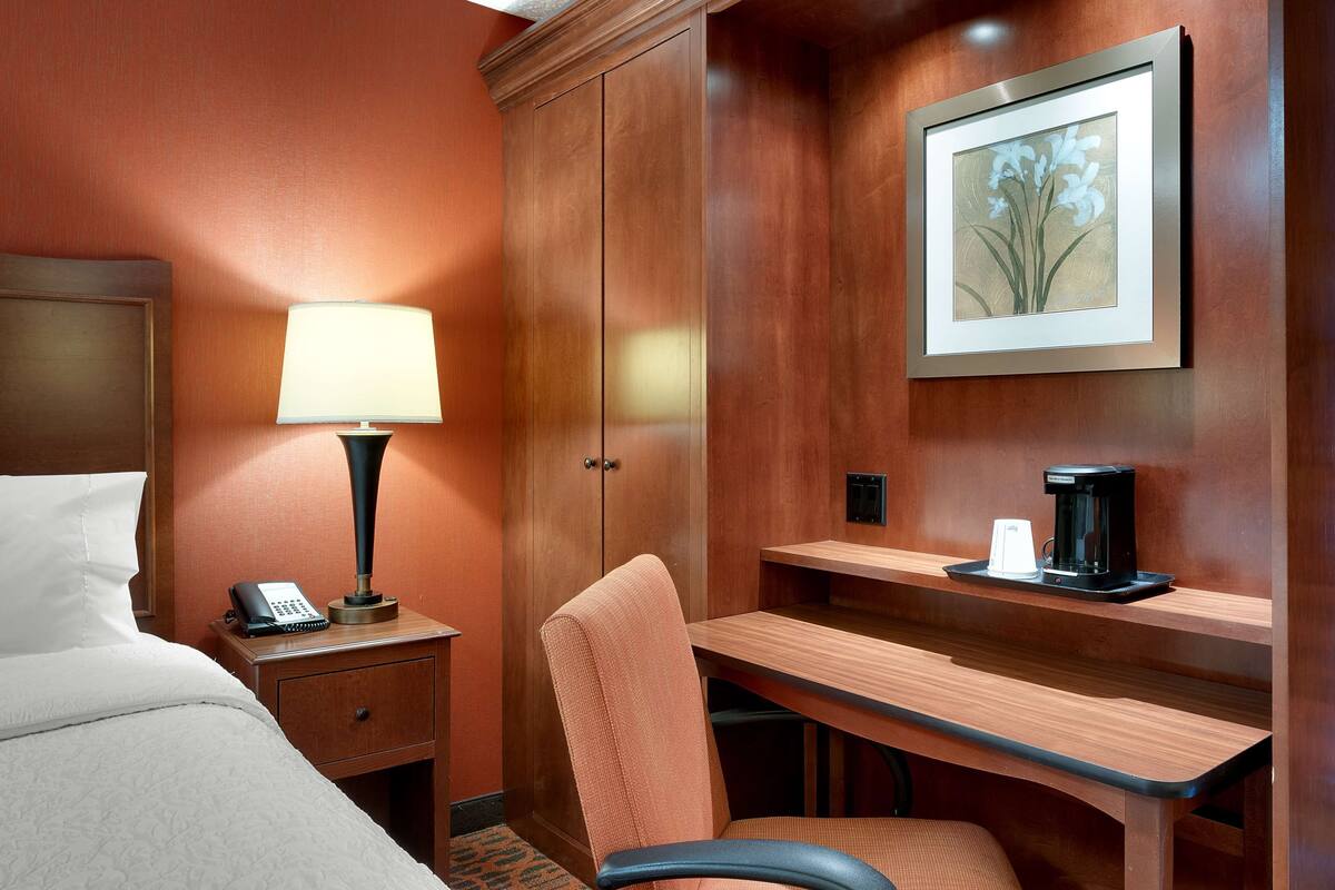 A hotel room with a bed, nightstand, lamp, desk, chair, and a framed picture on the wall.