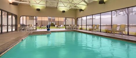 An indoor swimming pool with clear glass walls, a high ceiling with metal beams, and several lounge chairs around the pool.