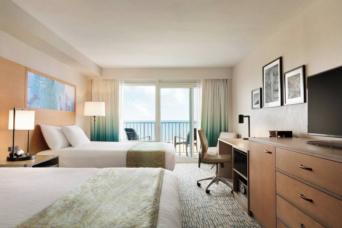 A hotel room with two beds, a desk, a chair, a TV, and a balcony with a view.