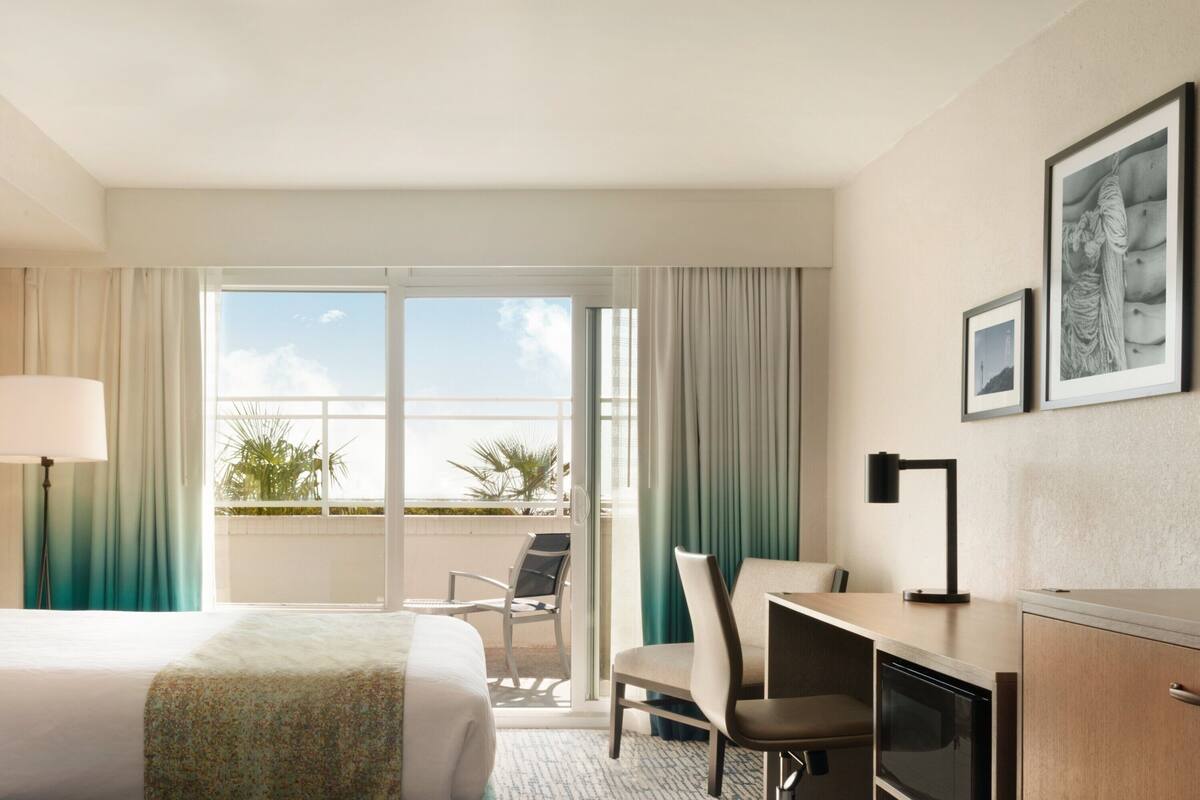 A hotel room with a bed, desk, chair, and a view through the glass door.