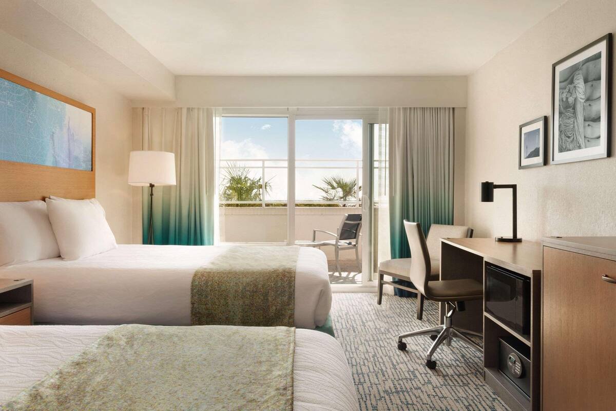 A hotel room with two beds, a desk, a chair, a lamp, and a view of the outdoors.