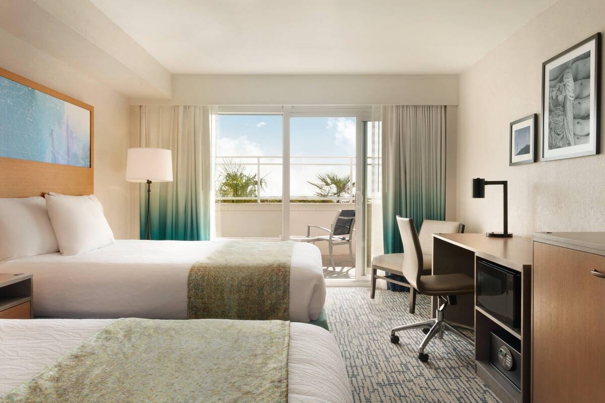 A hotel room with two beds, a desk, a chair, a lamp, and a view of the outdoors.