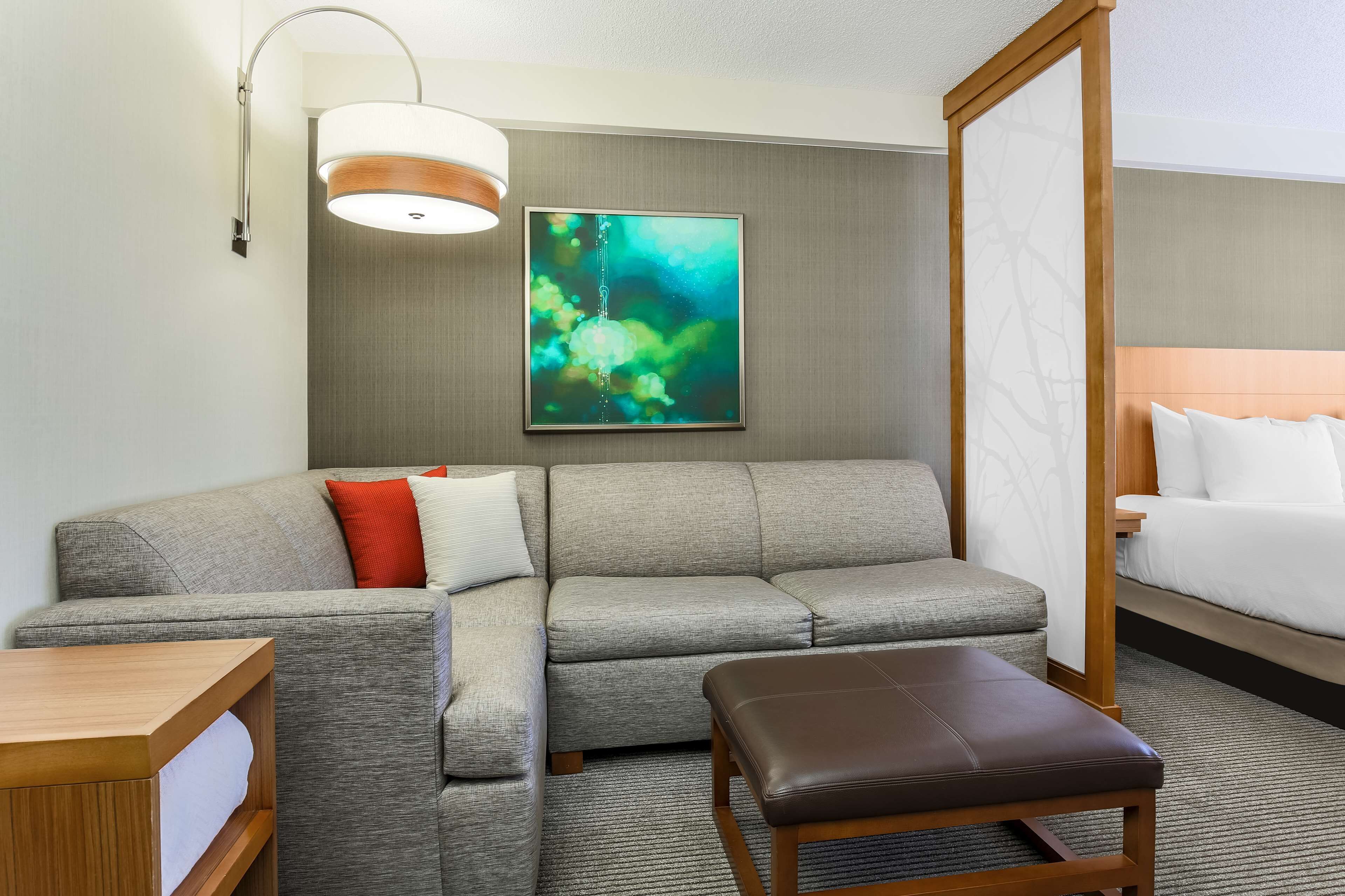 Une chambre d’hôtel avec un canapé gris, un banc en bois, une table de chevet, un grand lit, une œuvre d’art accrochée au mur et un luminaire au plafond.