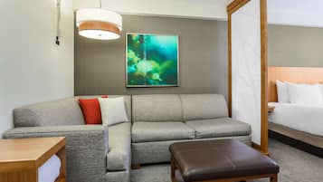 A hotel room with a grey sofa, a wooden bench, a bedside table, a large bed, a wall-mounted artwork, and a ceiling light.