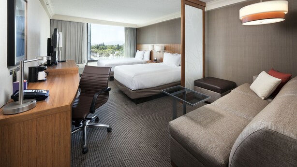 Quarto de hotel com cama, escrivaninha com computador, cadeira, sofá e uma ampla janela com vista.