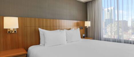 Habitación de hotel con una cama grande, cabecera de madera, dos mesitas de noche con lámparas y un ventanal con vistas a la ciudad.