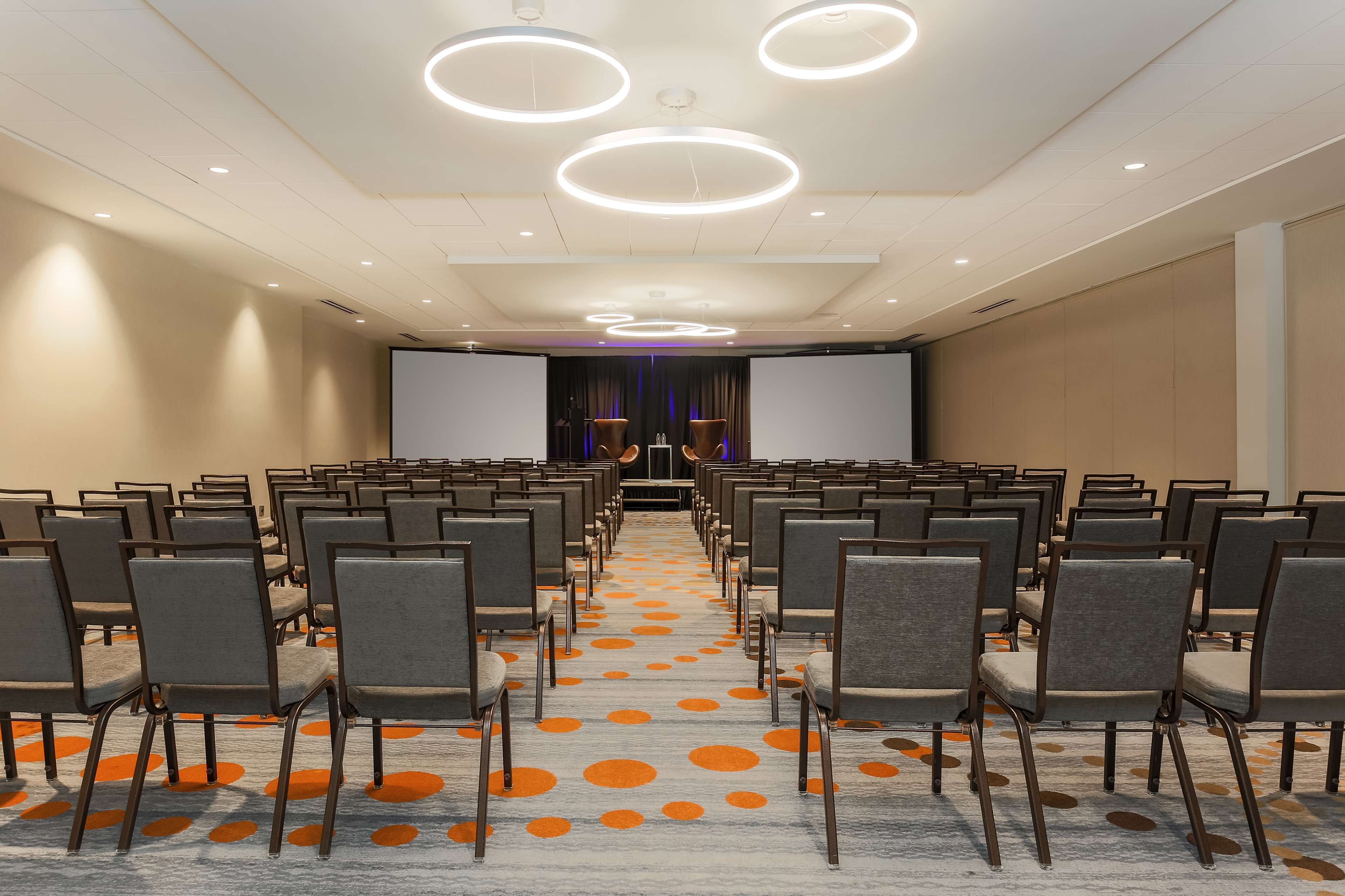 A conference room with rows of chairs, a large screen at the front, and circular ceiling lights.