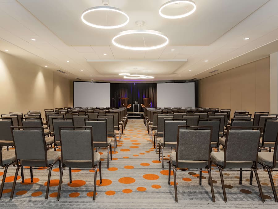 A conference room with rows of chairs, a large screen at the front, and circular ceiling lights.