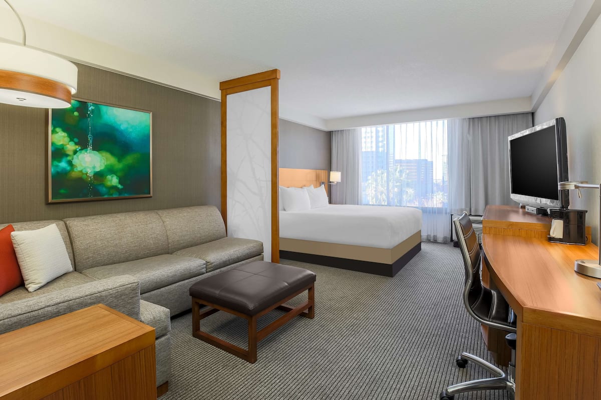 Ett hotellrum med en soffa, en säng, ett skrivbord med en dator, en tv och ett stort fönster med gardiner.