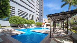 Una piscina con sillas de descanso y una zona de estar cubierta, rodeada de vegetación y edificios modernos.