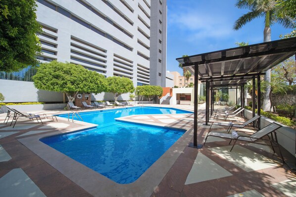 A swimming pool with lounge chairs and a covered seating area, surrounded by greenery and modern buildings.