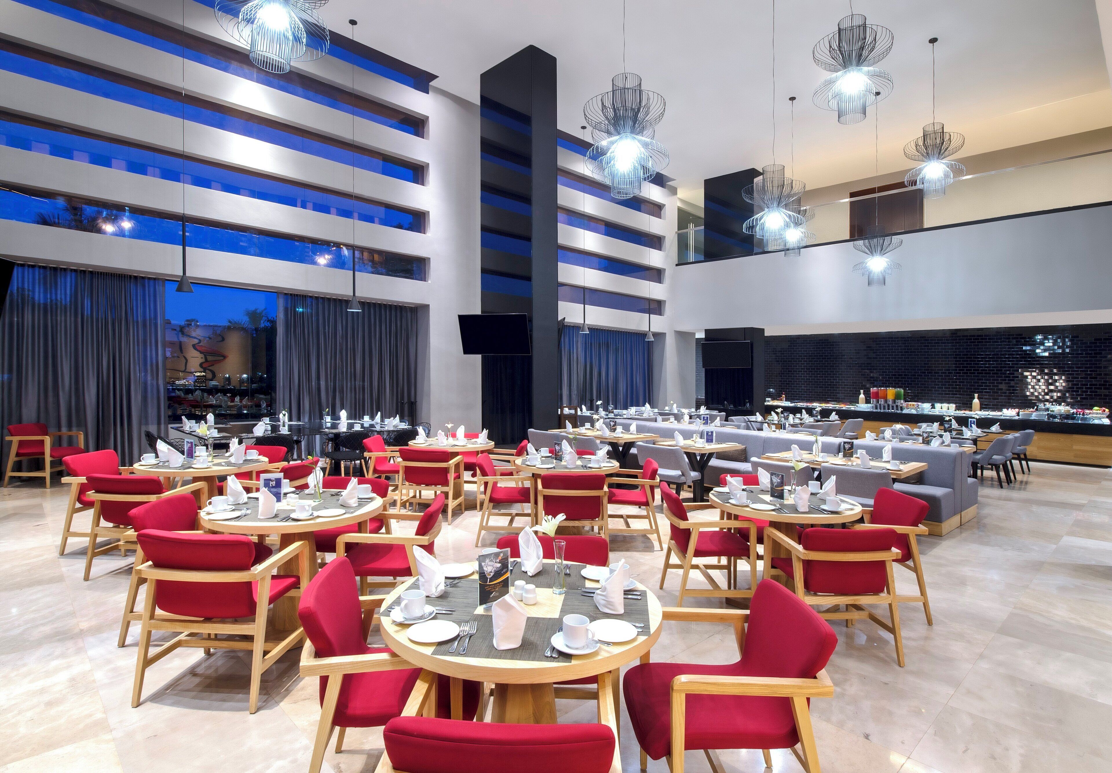 A modern restaurant with round tables and red chairs, large windows, and contemporary lighting fixtures.