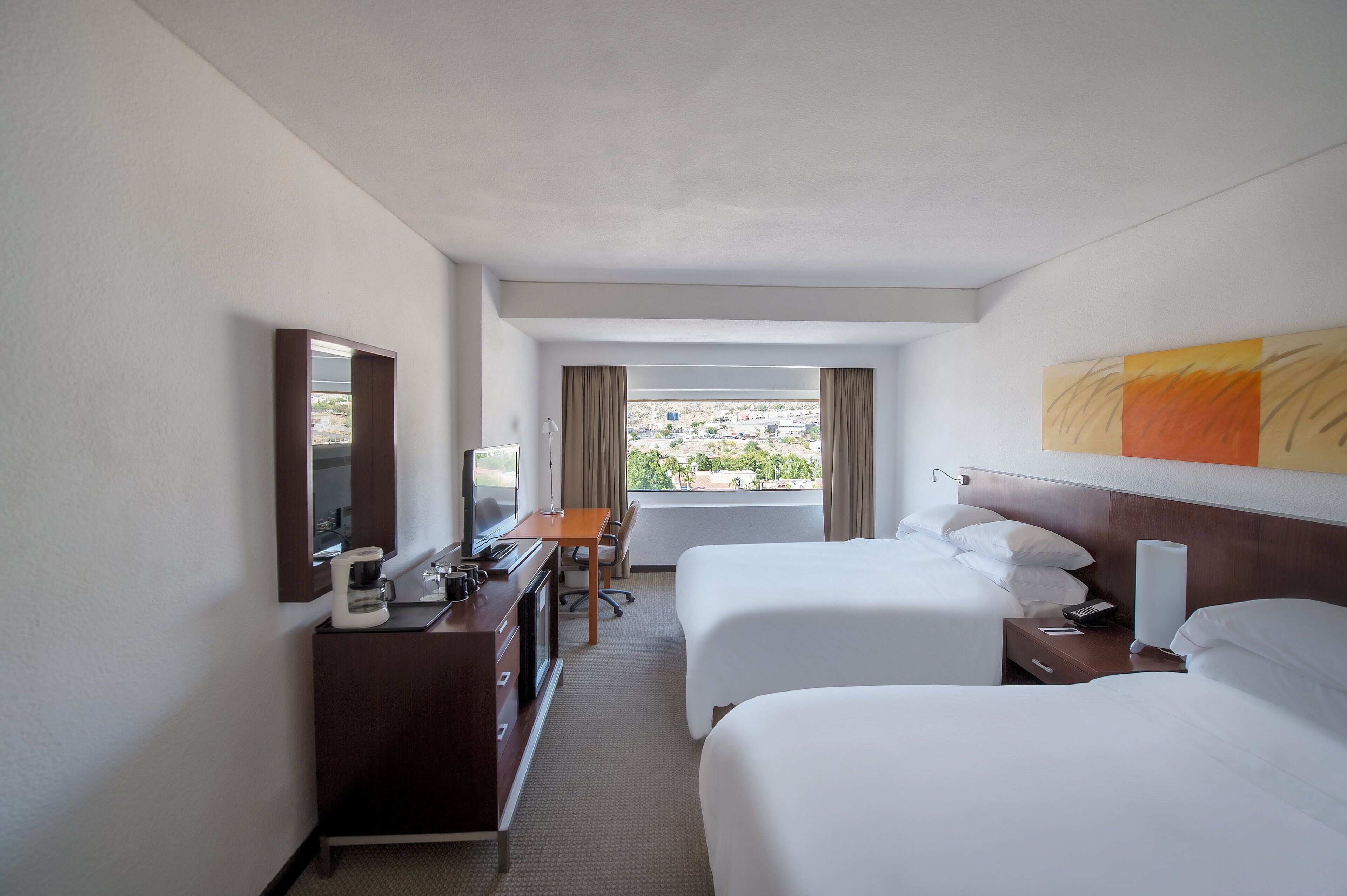 Une chambre d’hôtel avec deux lits, un bureau avec une chaise, une commode, un miroir, une fenêtre avec des rideaux et un tableau au mur.