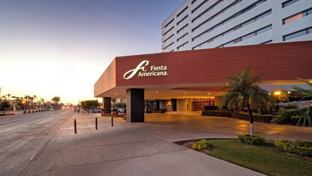 Una entrada de hotel moderna con un llamativo letrero 'Fiesta Americana', amplias aceras y un área exterior bien cuidada con palmeras.