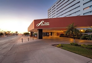 A modern hotel entrance with a prominent 'Fiesta Americana' sign, wide pedestrian pathways, and a well-maintained outdoor area with palm trees.