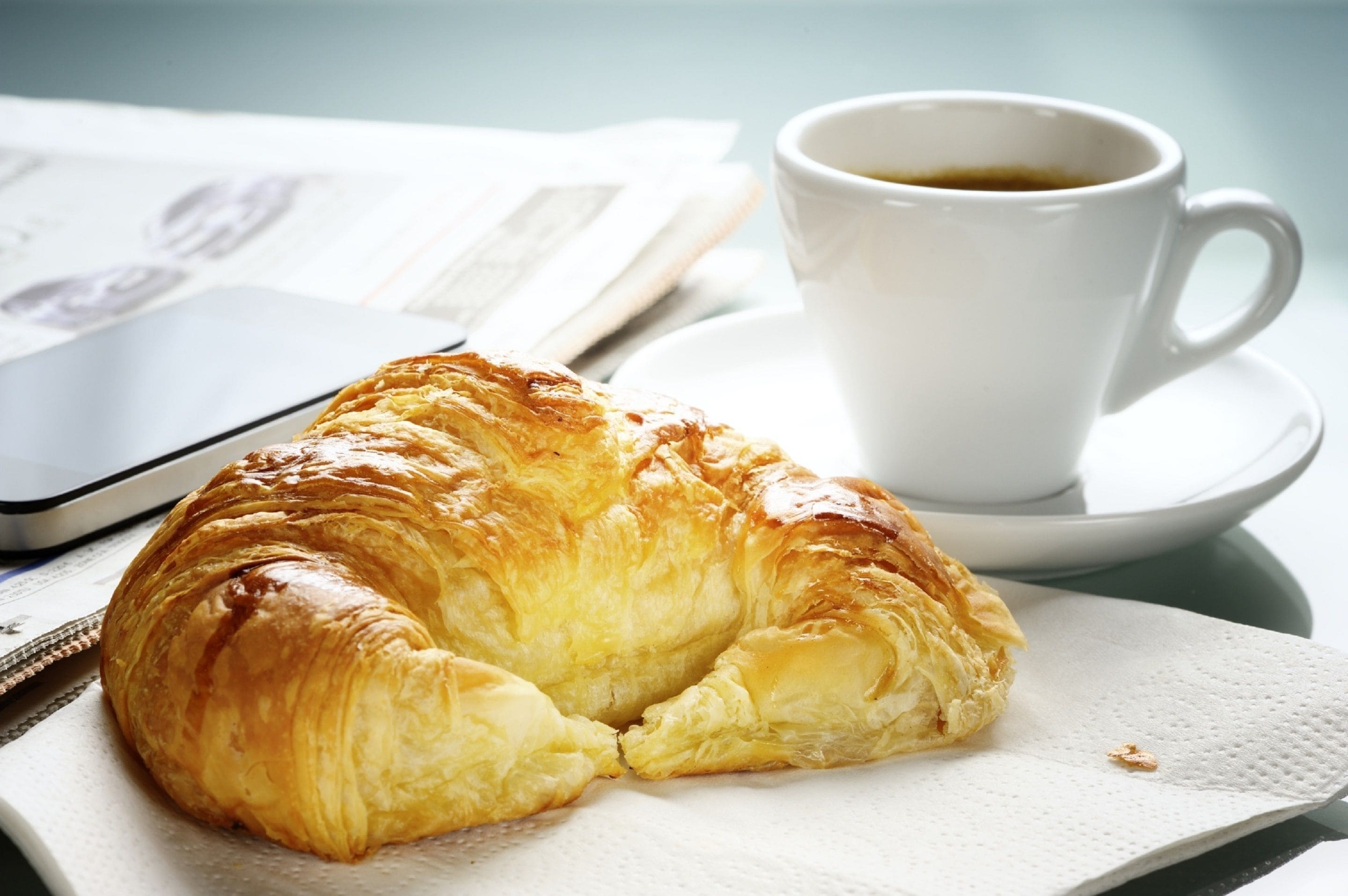 A croissant on a napkin, a cup of coffee, and a smartphone on a table.