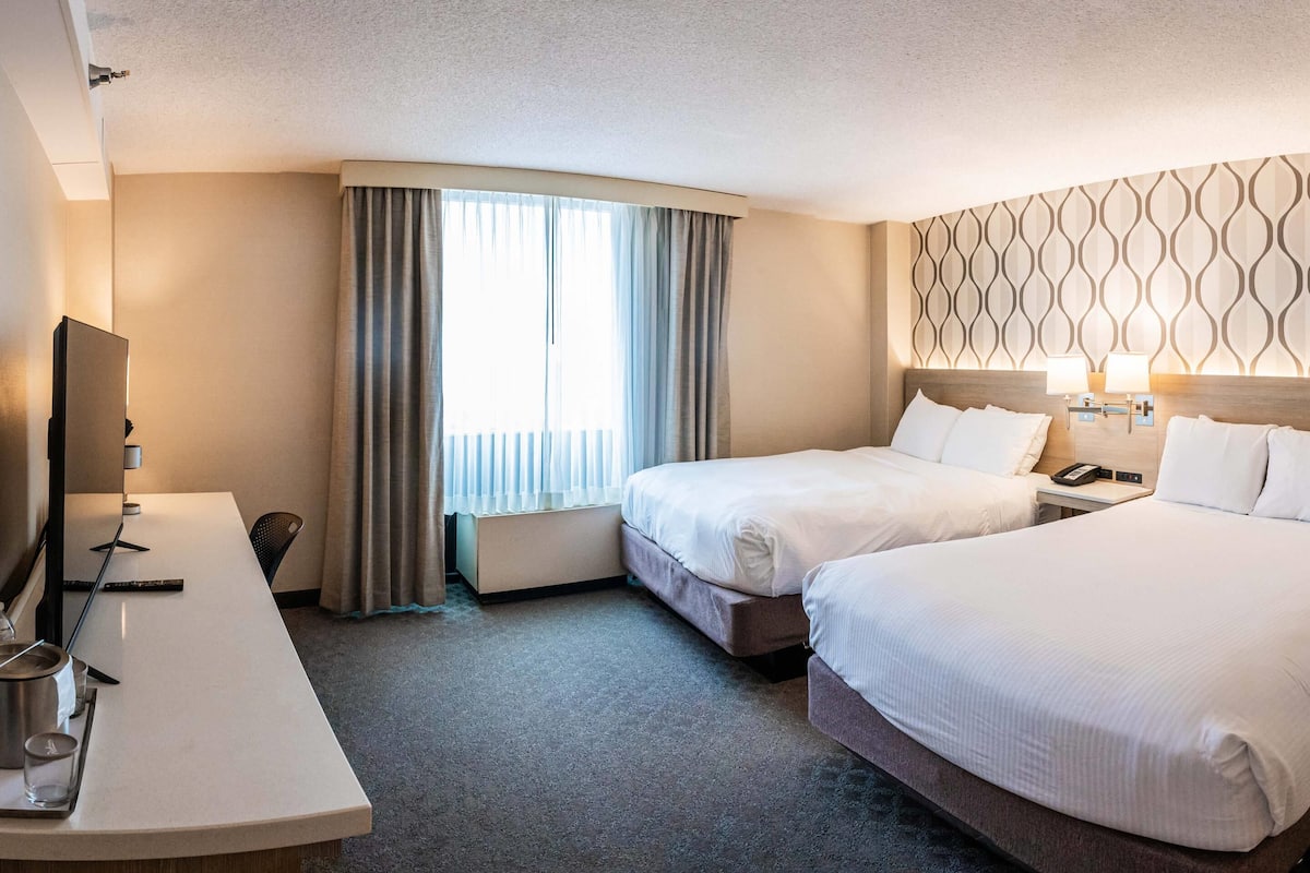 A hotel room with two beds, a desk, a TV, and a window with curtains.