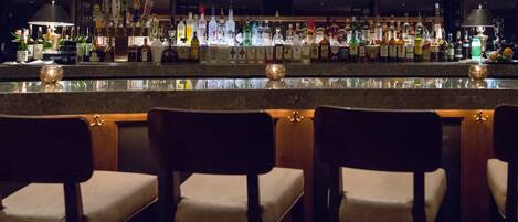A bar area with multiple liquor bottles, a well-lit bar counter, and several chairs.