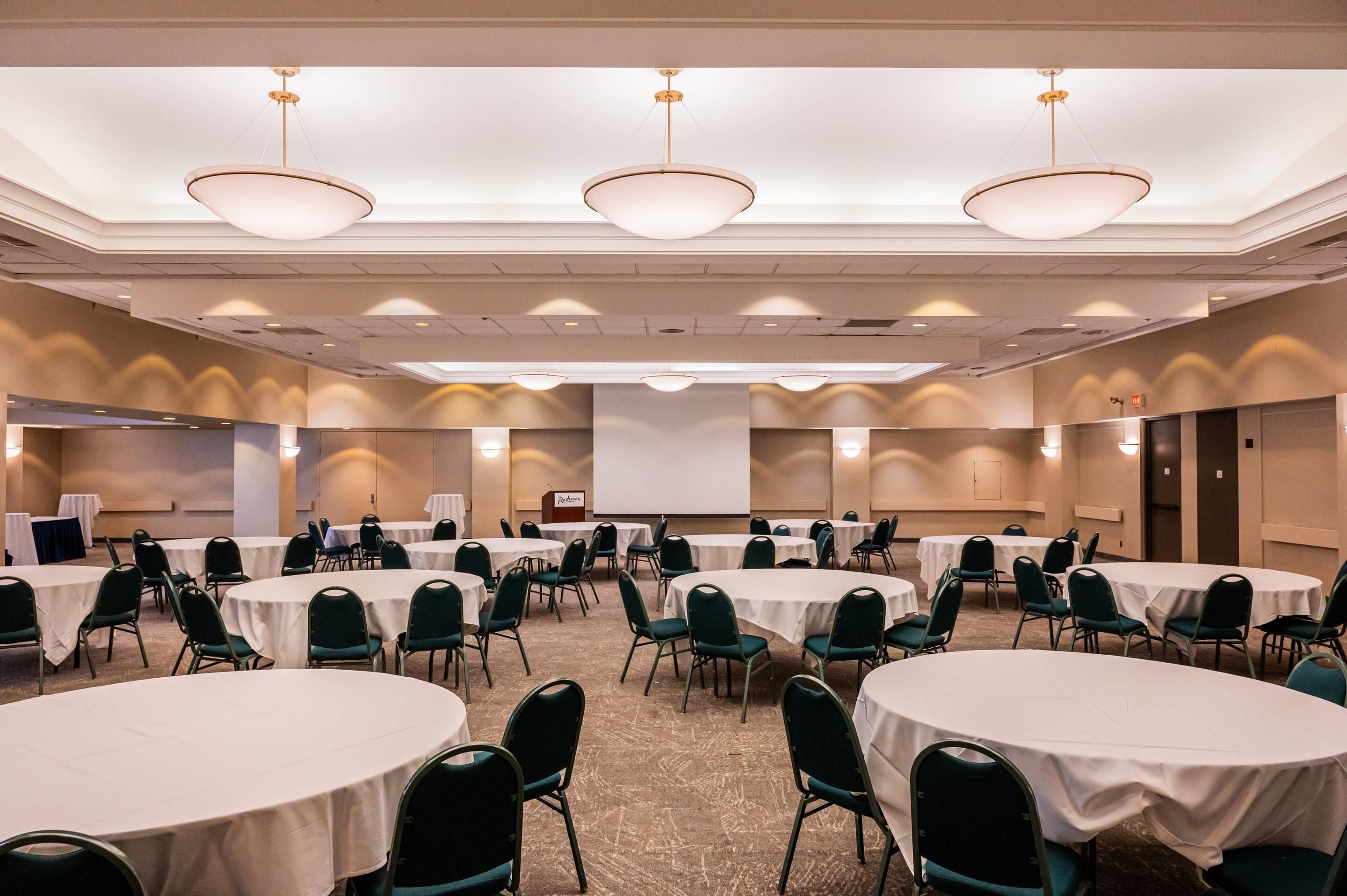 A conference room with multiple round tables, green chairs, and a large screen at the front.
