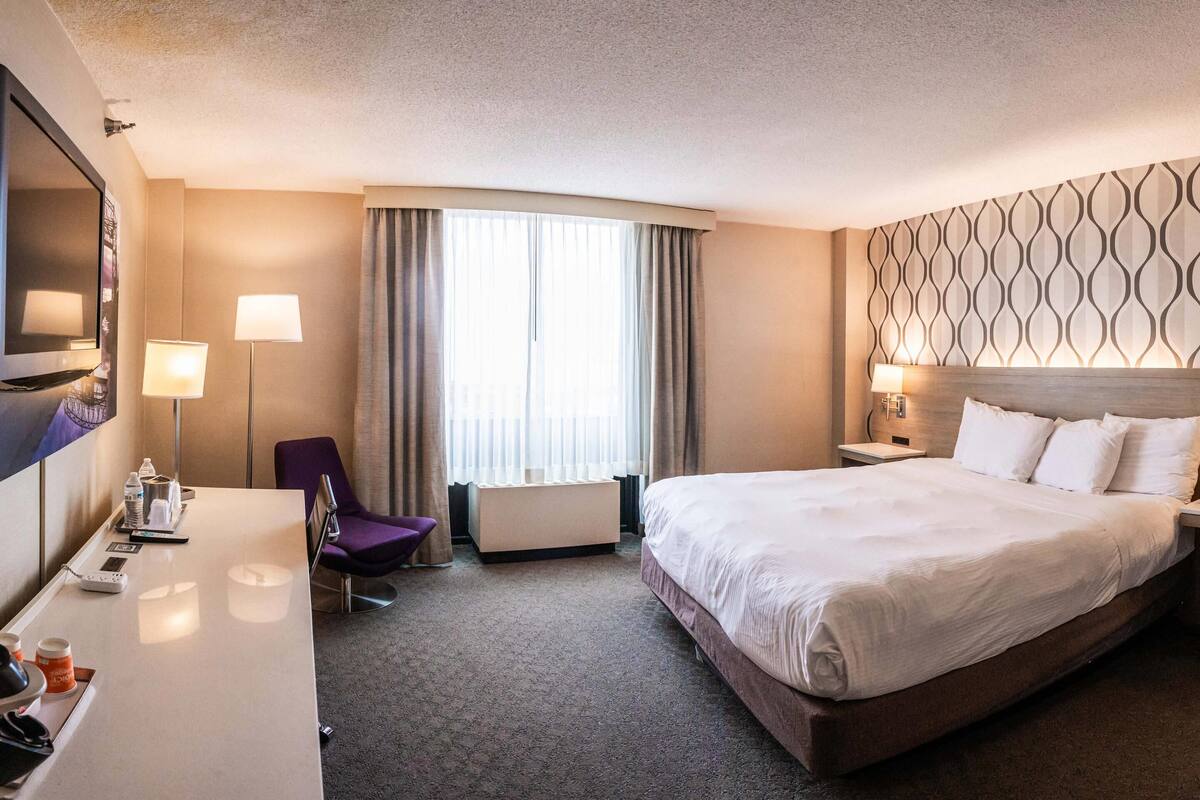 A hotel room with a large bed, a desk with a chair, a flat-screen TV, a bedside table with a lamp, and a window with curtains.