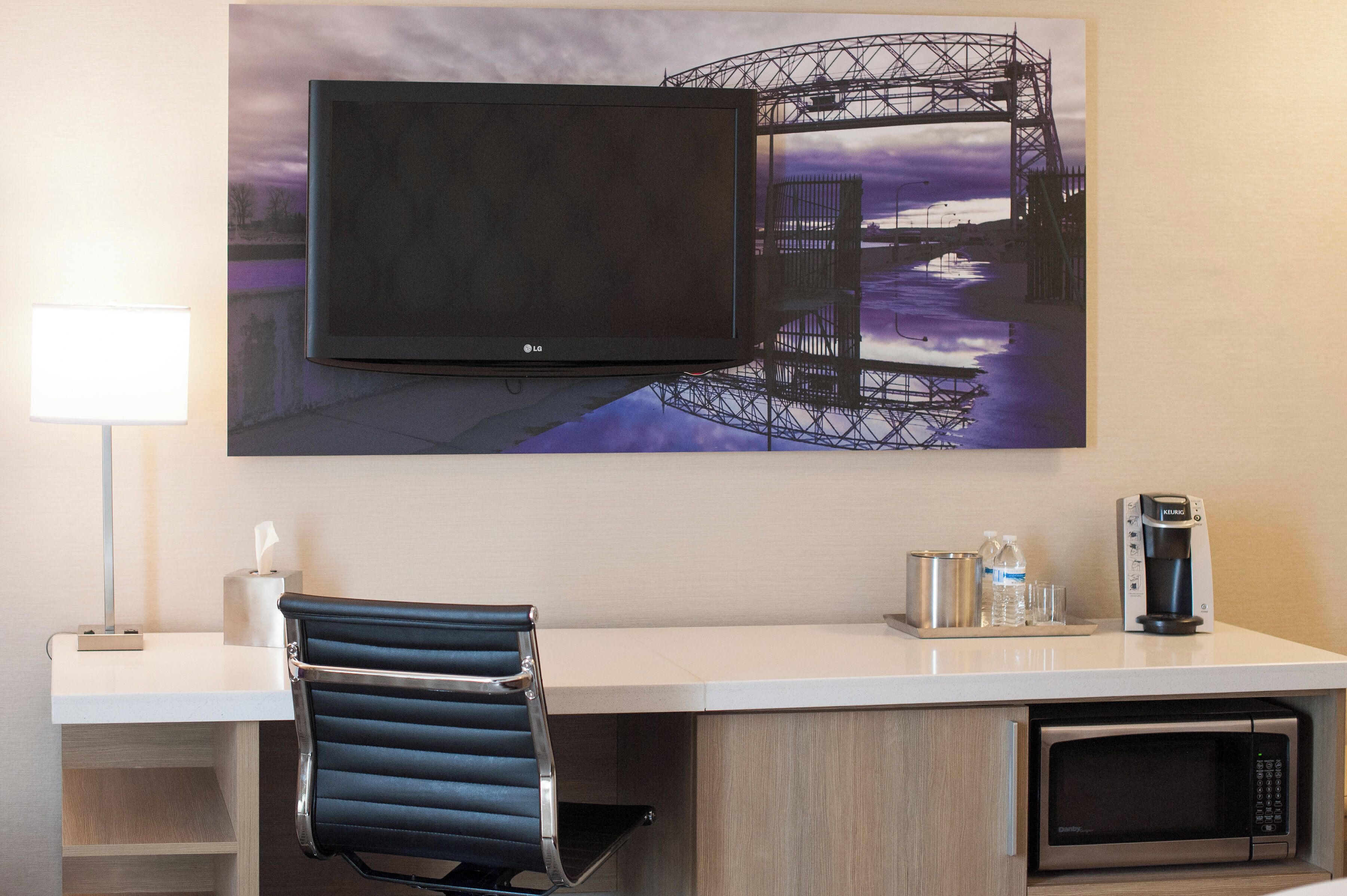 A hotel room with a flat-screen TV mounted on the wall, a desk with a chair, a microwave, a coffee maker, and a water dispenser.